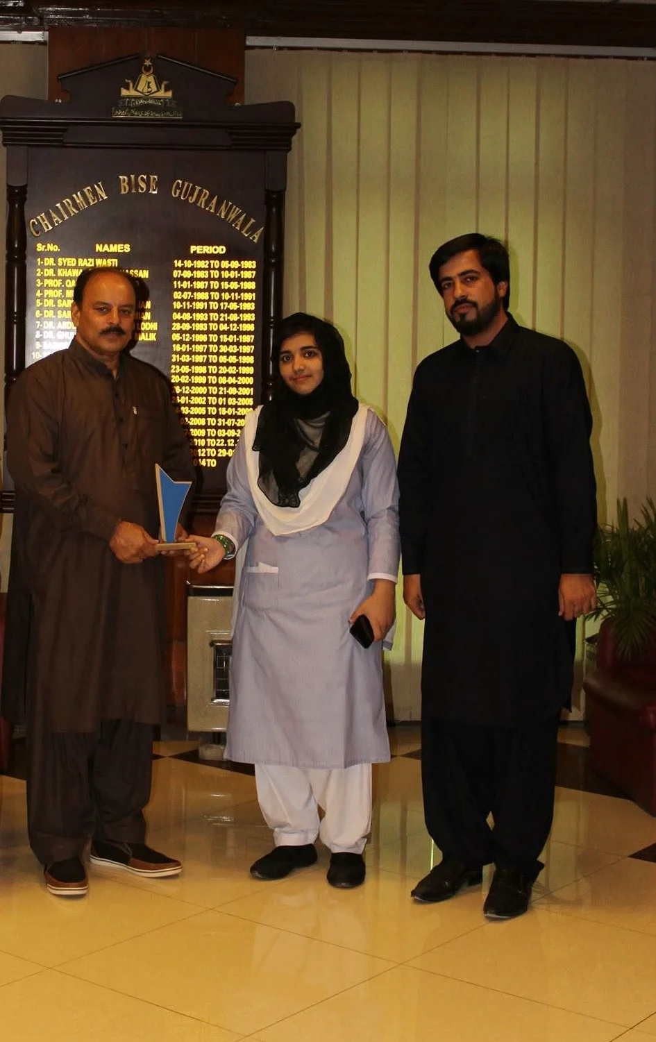 A Danish International Girls College student receiving a commemorative shield from the BISE Gujranwala Chairman, accompanied by Faruukh Nadeem.